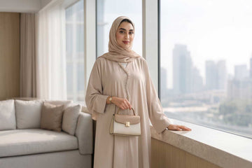 Woman in a taupe open-front abaya with matching hijab holding a structured handbag by a window overlooking the Dubai skyline, illustrating everyday luxury abaya styling.