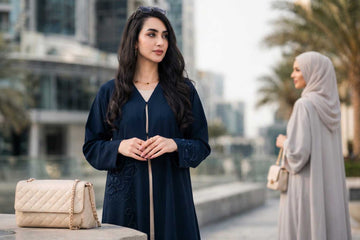 Woman in modern navy abaya with tonal embroidery styled with quilted handbag beside a second woman in warm neutral abaya in Dubai - Layaal