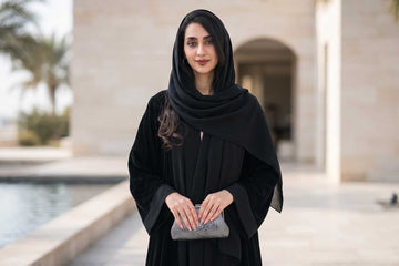 Woman in timeless black abaya and matching black hijab holding silver mesh clutch by a reflecting pool in a luxury Mediterranean setting - Layaal