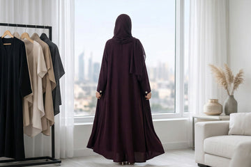 Woman in custom fit burgundy open abaya with matching sheila standing by floor to ceiling window overlooking Dubai at Layaal Abaya Studio